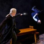 Draco and Harry fighting. Draco holds his wand out whihc is lit up. Harry is suspended in the air, upside down!