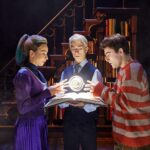 Delphi Diggory (left), Scorpius Malfoy (centre) and Albus Potter (right) stand around a floating time turner with a book underneath. They are wearing the clothes we see Hermione, Harry and Ron wearing. Behind is a staircase with a bookcase underneath