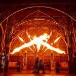 On stage production shot. A church-like setting is lit up with flames. The cast of wizards all point their wands to the centre of the stage.